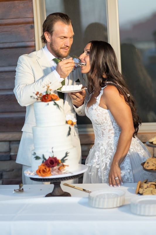 Bride and groom cutting and sharing wedding cake outdoors, dressed in white wedding attire at their reception.