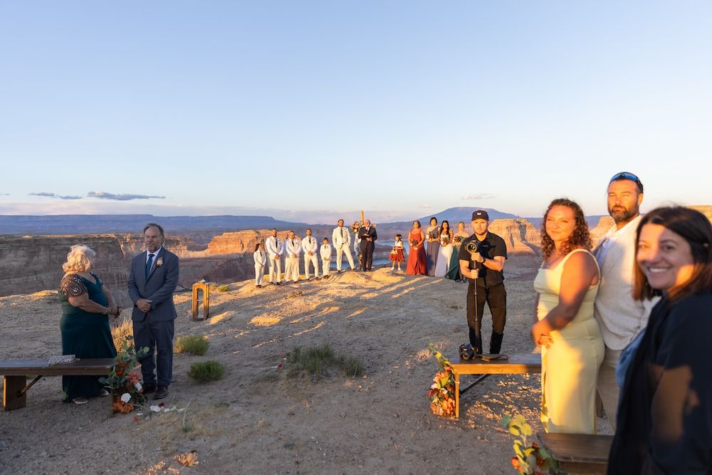 Wedding ceremony at sunset on desert canyon overlook with guests gathered around couple exchanging vows.