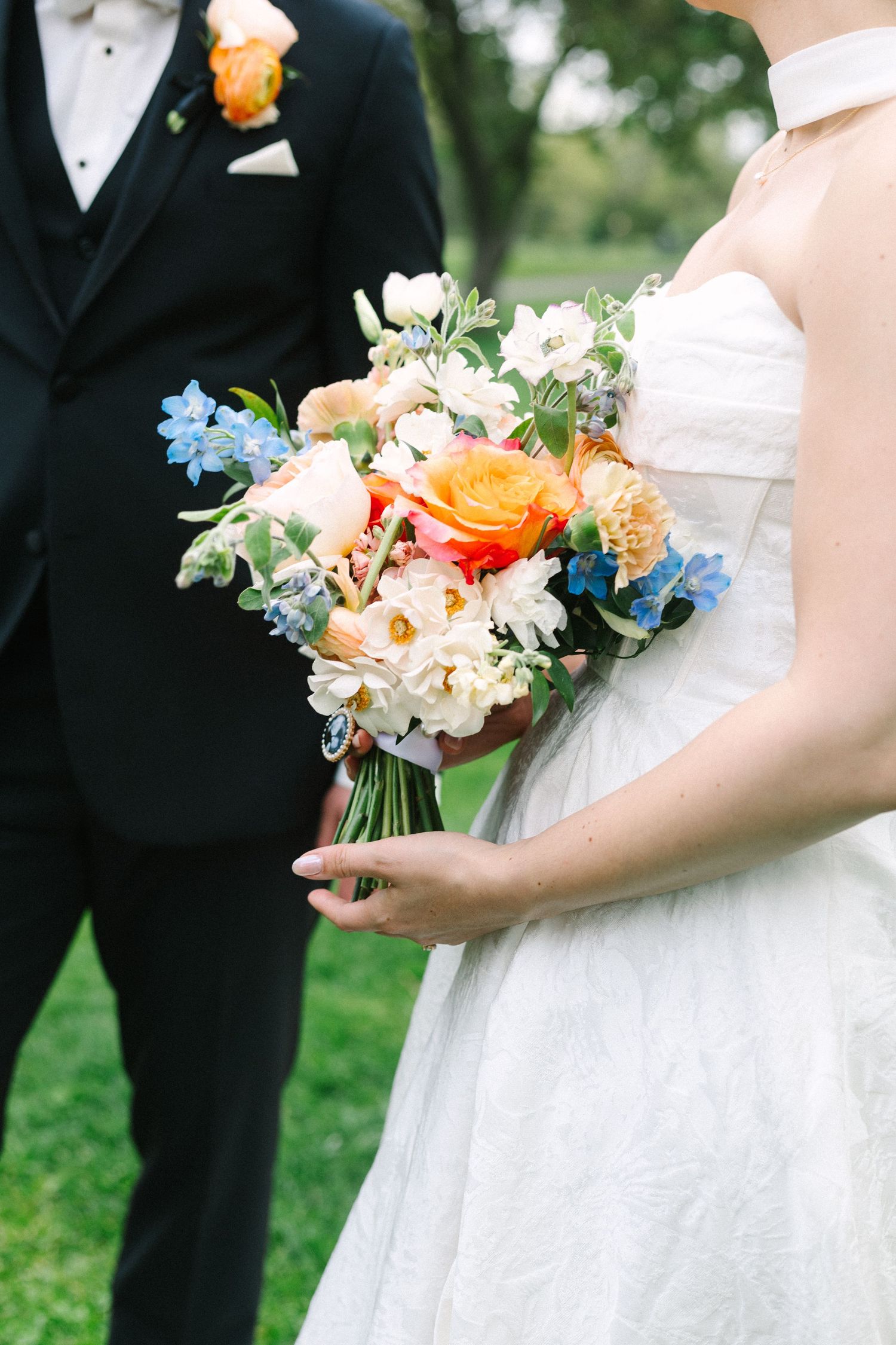 Bride holding colorful wedding bouquet with orange, blue, and white flowers beside groom in black tuxedo.