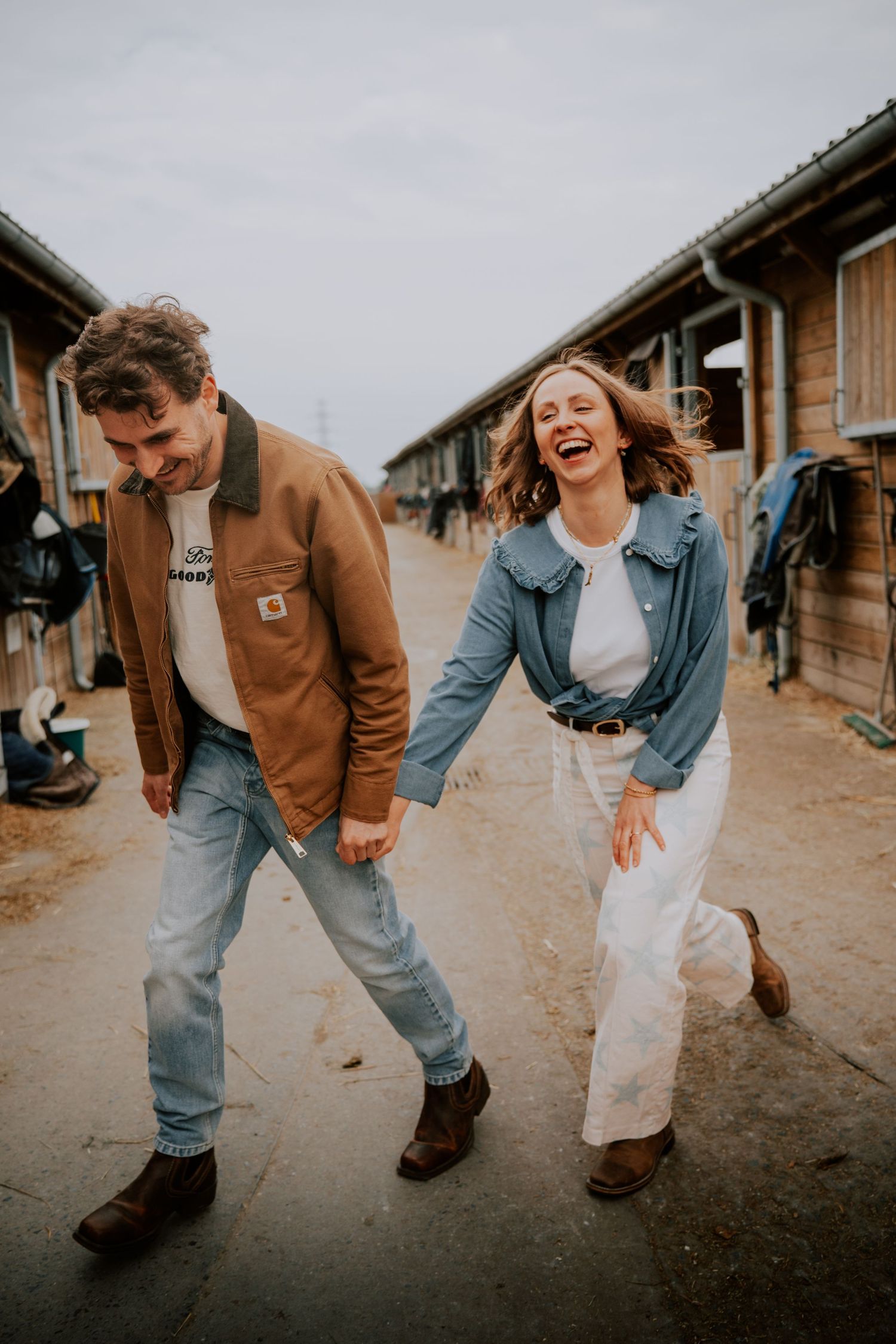 A laughing couple walks hand-in-hand past wooden stables, wearing casual denim outfits and brown boots on a cloudy day.