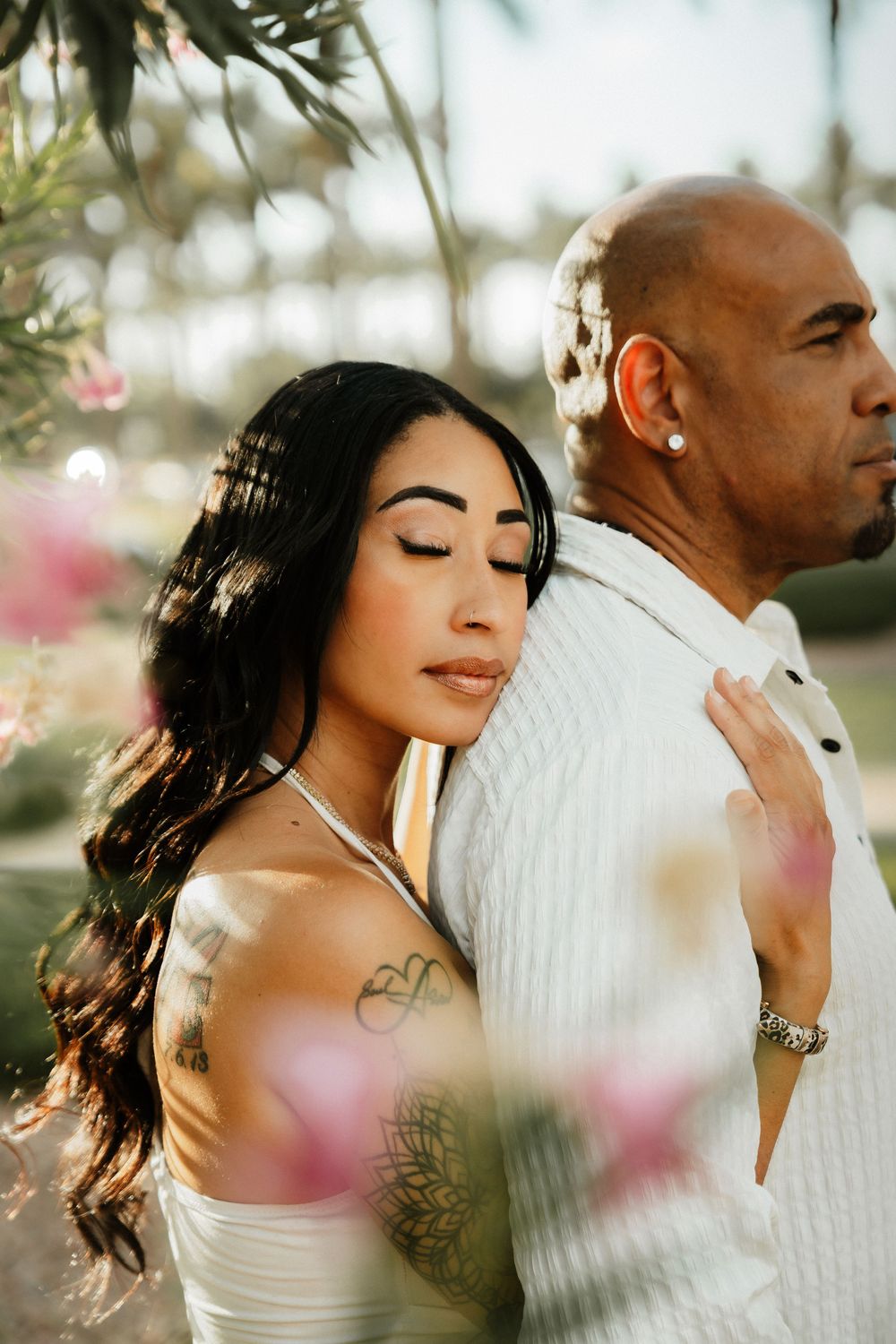 A tattooed bride leans against a bald groom in white attire, surrounded by pink flowers and palm trees in soft sunlight.