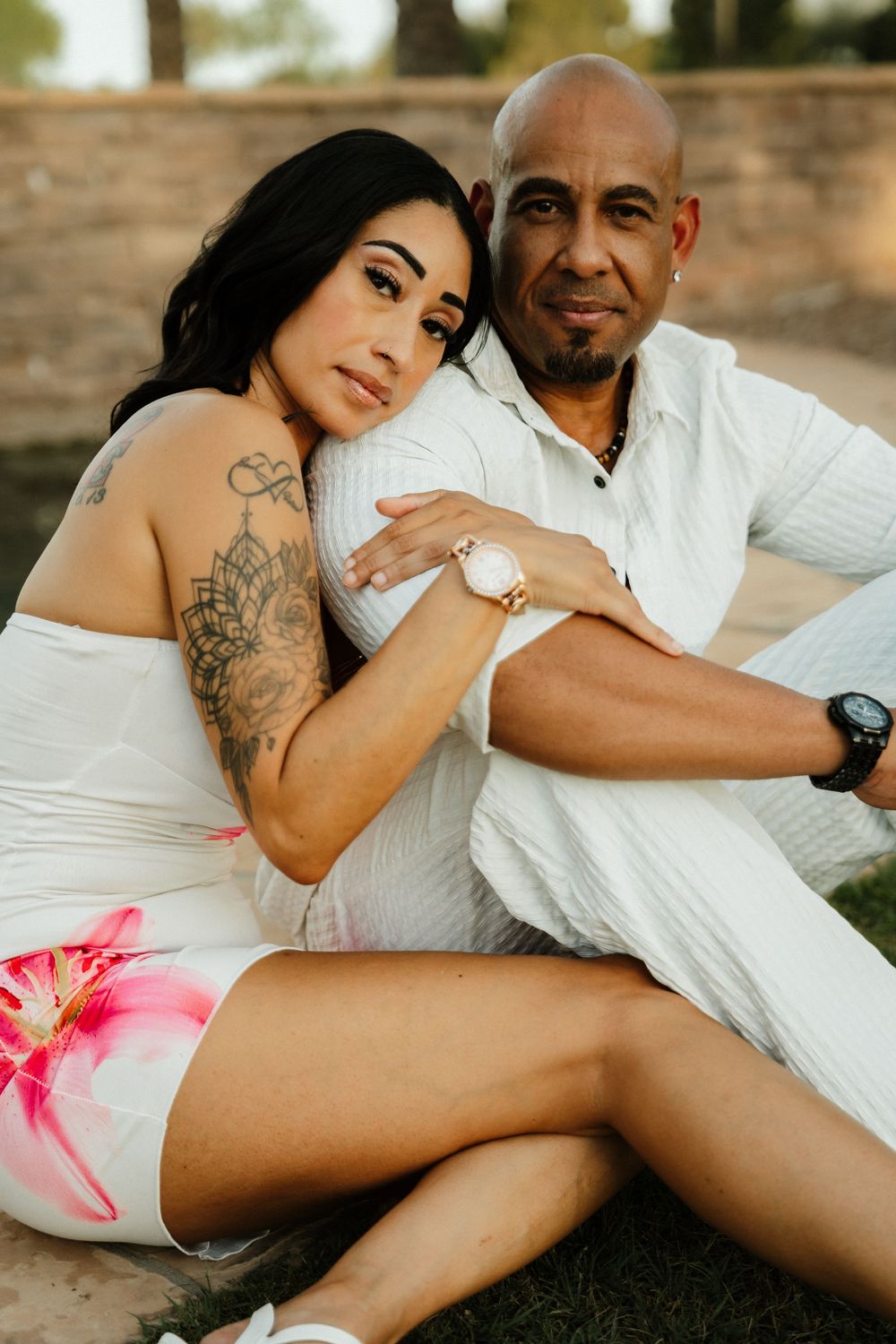 A couple in white outfits sit together on grass, woman with tattoos leans on man's shoulder in warm outdoor light.