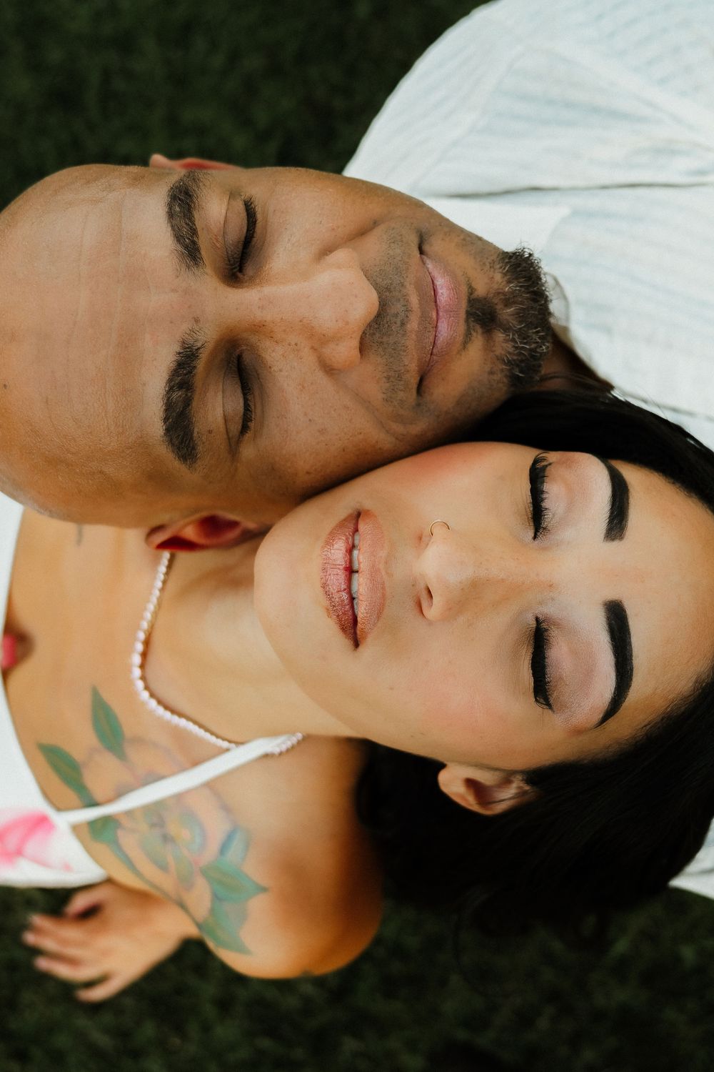 Overhead view of a smiling couple lying together on grass, woman wearing pearl necklace with floral tattoos.