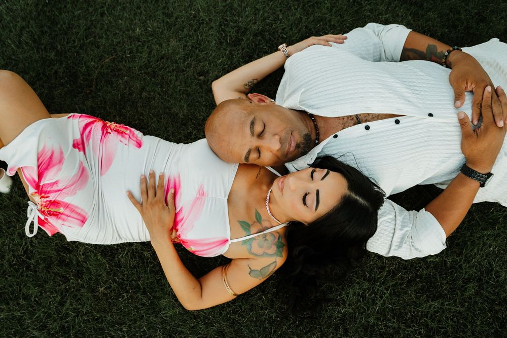Couple lying on grass in romantic pose, woman in floral dress with tattoos, man in white shirt, eyes closed.