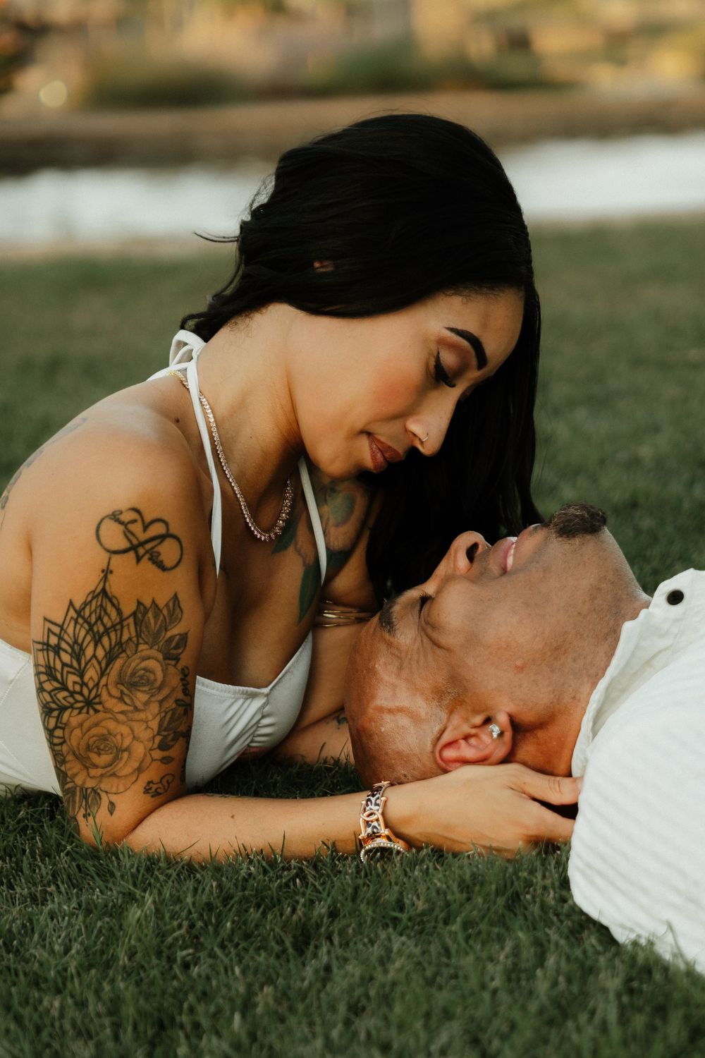 A tattooed woman in a white top leans over a man lying on grass in an intimate romantic outdoor moment.