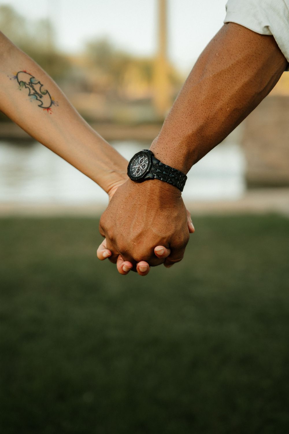 Two people holding hands outdoors, one with a floral tattoo and the other wearing a black watch.
