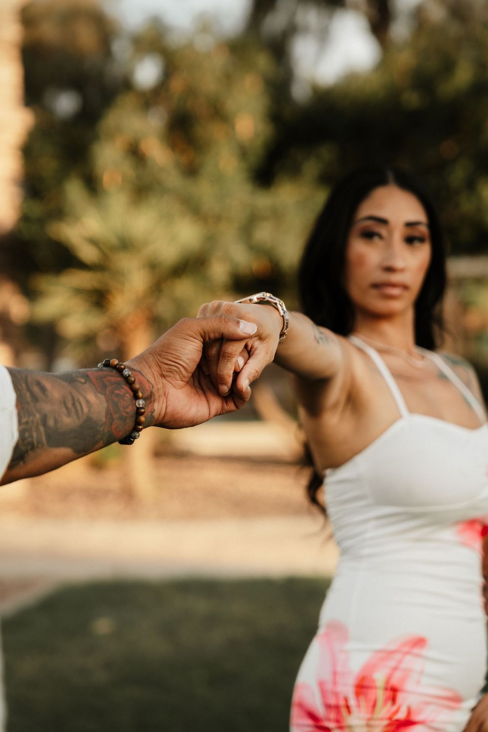 A tattooed man holds a woman's hand in a sunny park, she wears a white floral dress.