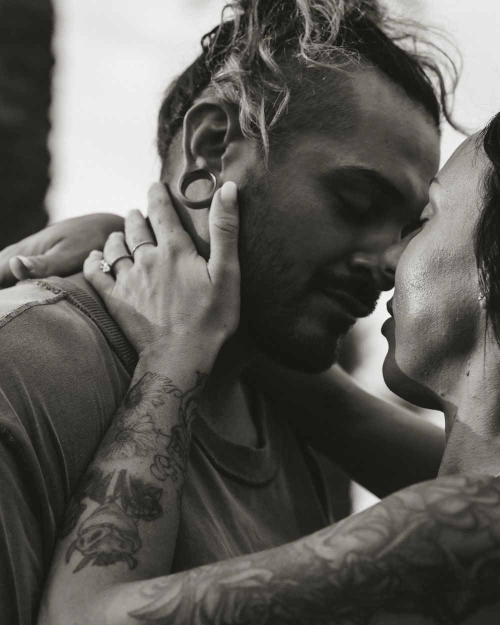 Black and white photo of tattooed couple sharing an intimate moment, man with gauged ears and curly hair tenderly touching womans face.