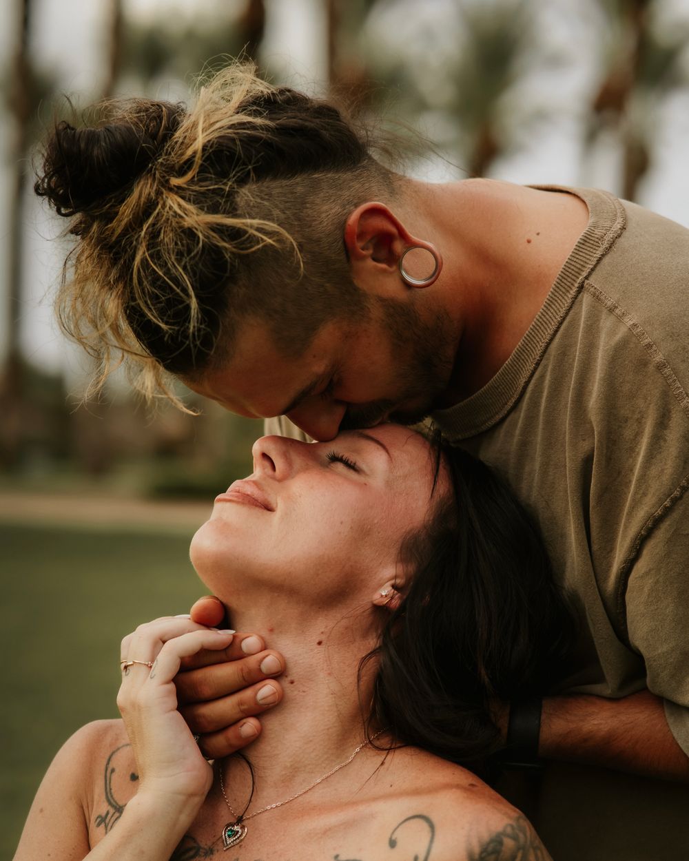 A couple shares an intimate moment, man with man bun and gauge earrings kissing woman with tattoos on her neck outdoors.