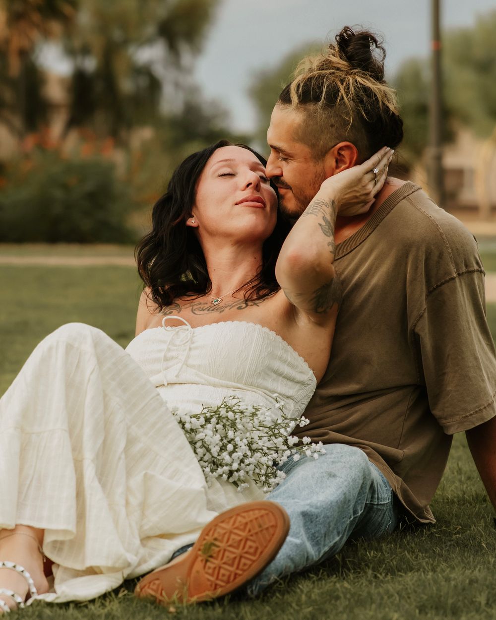 Couple sitting on grass, woman in white dress holding baby's breath flowers while man tenderly kisses her cheek outdoors.