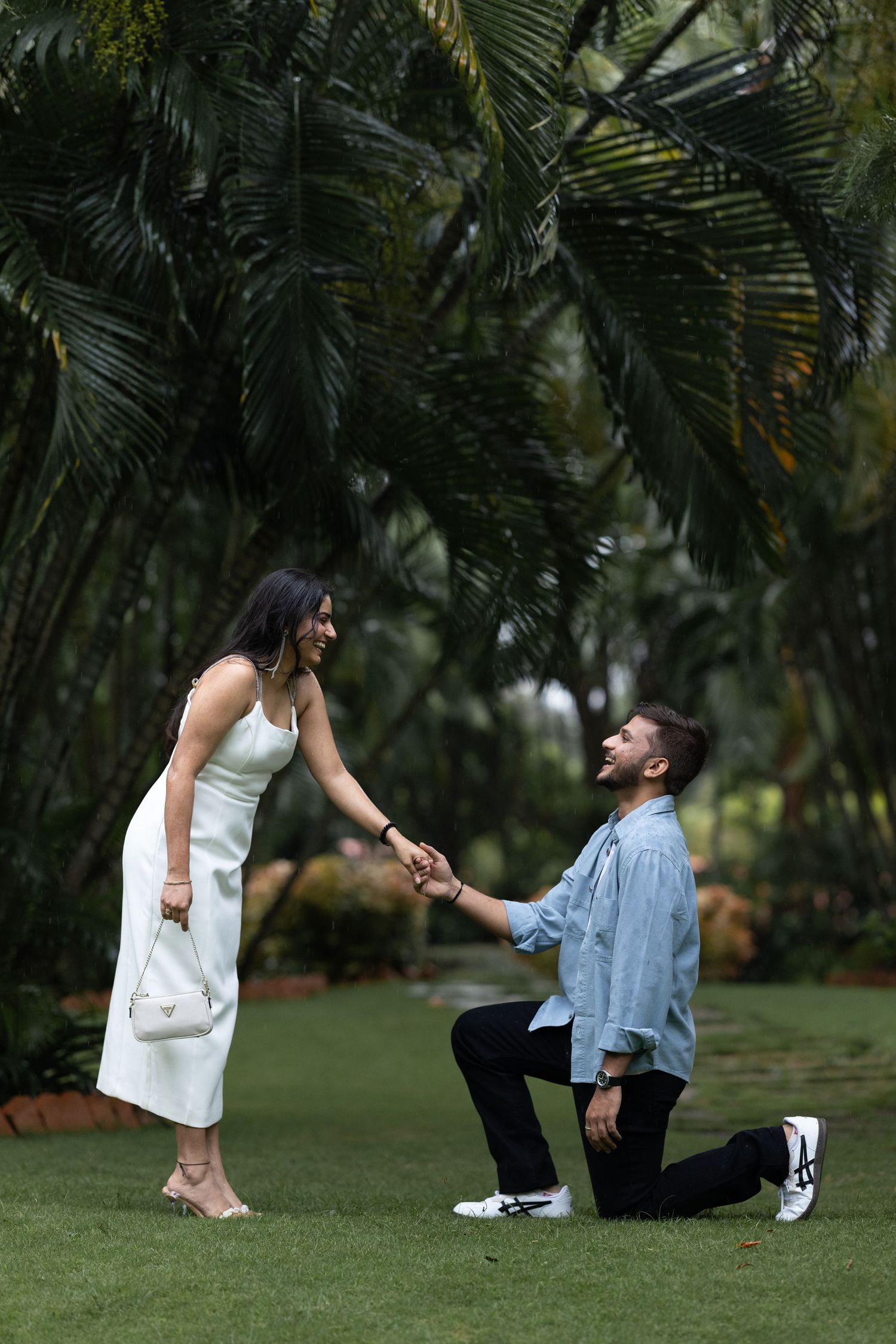Man on one knee proposing to a woman in a white dress in a lush tropical garden setting.