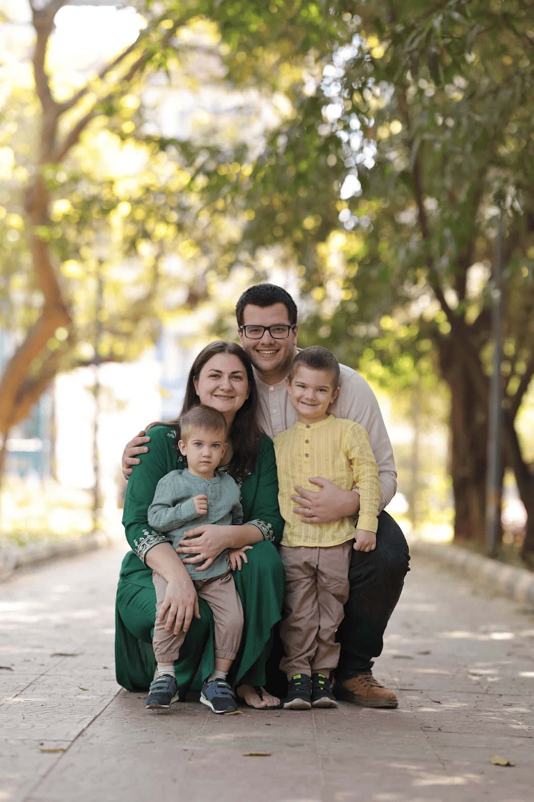 Happy family of four posing outdoors in a tree-lined park, parents smiling with two young boys.