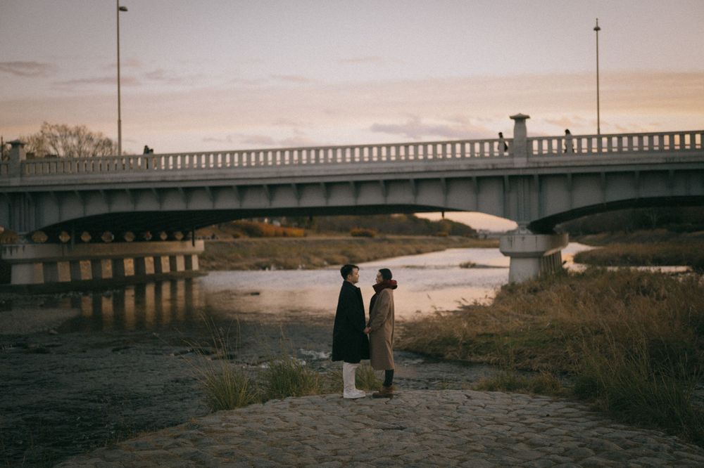 Couple standing by a river at sunset with a concrete bridge in the background.