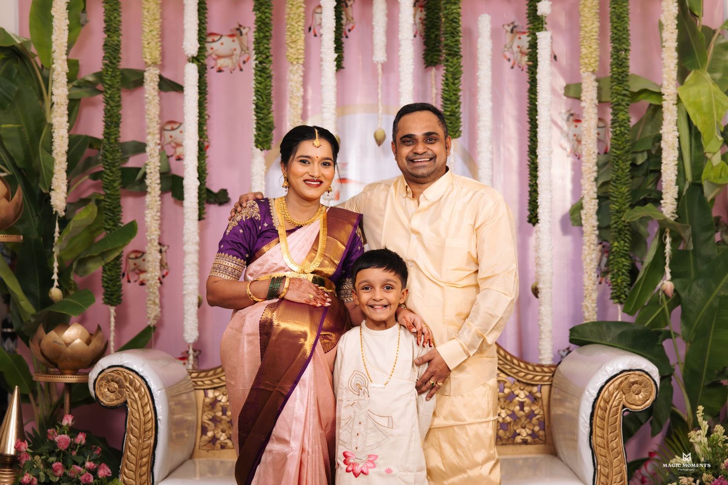 Family of three posing at a South Indian wedding celebration with floral backdrop and traditional silk attire.