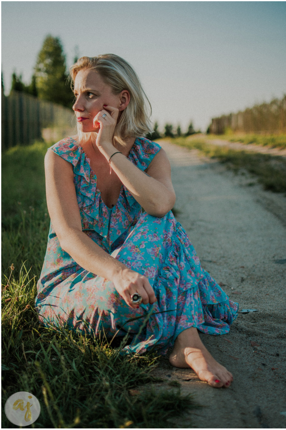 Blonde woman in blue floral dress sitting barefoot on dirt path, gazing thoughtfully into distance at golden hour.