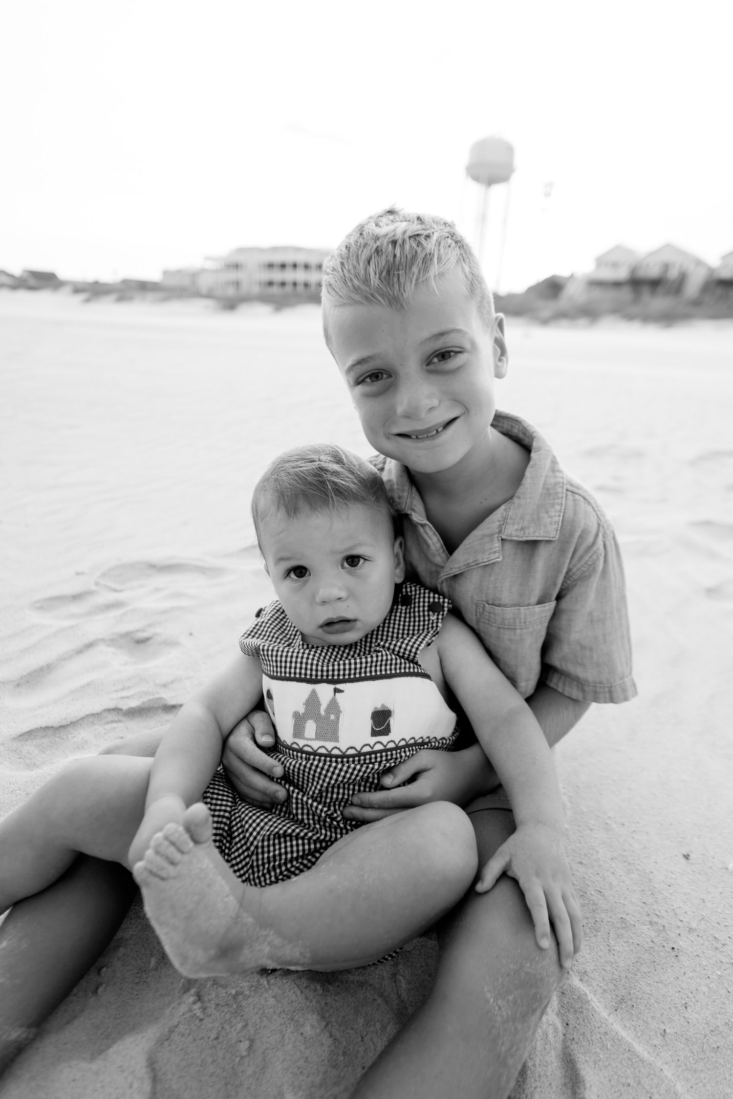 Black and white photo of older boy holding baby sibling on sandy beach, smiling at camera.