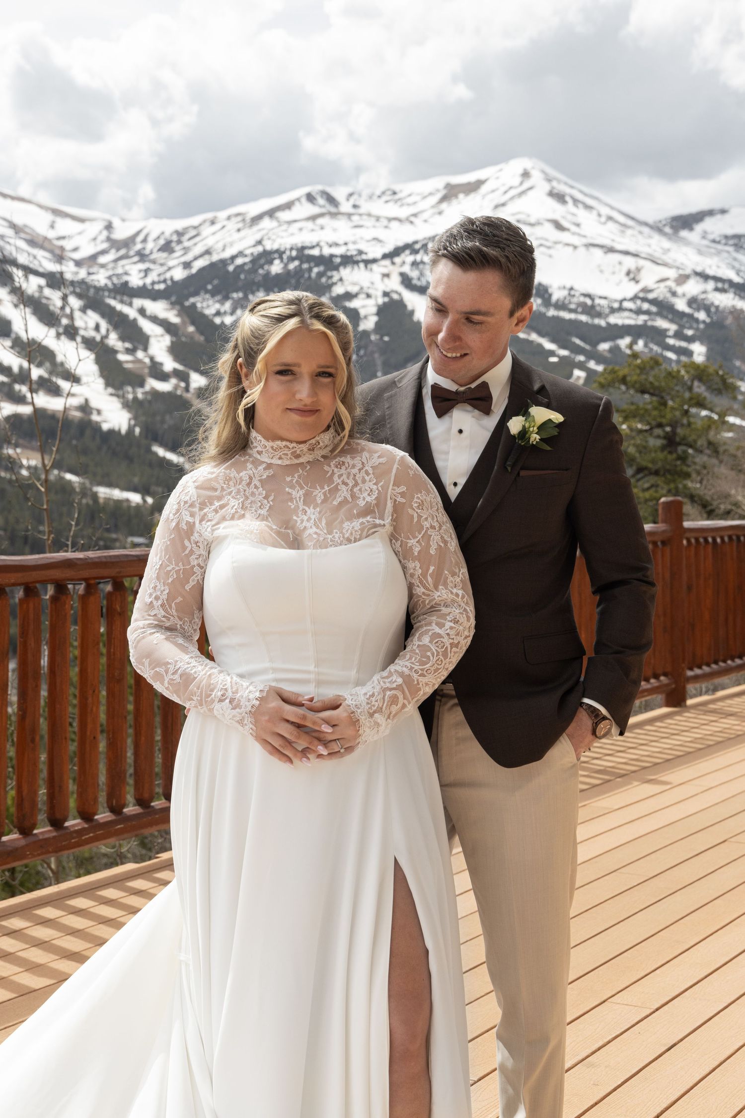 Black and white photo of bride in lace long-sleeve gown embraced by groom in tuxedo with bow tie.