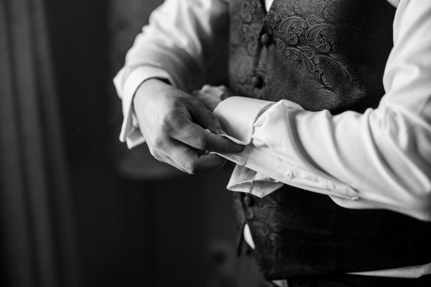 Black and white photo of a groom fastening his cufflinks while wearing a paisley vest and white dress shirt.