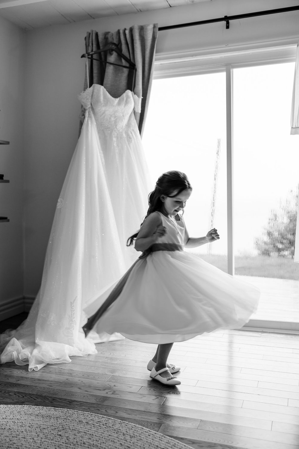 A flower girl twirls in a white dress beside a hanging wedding gown near a bright window, black and white photo.