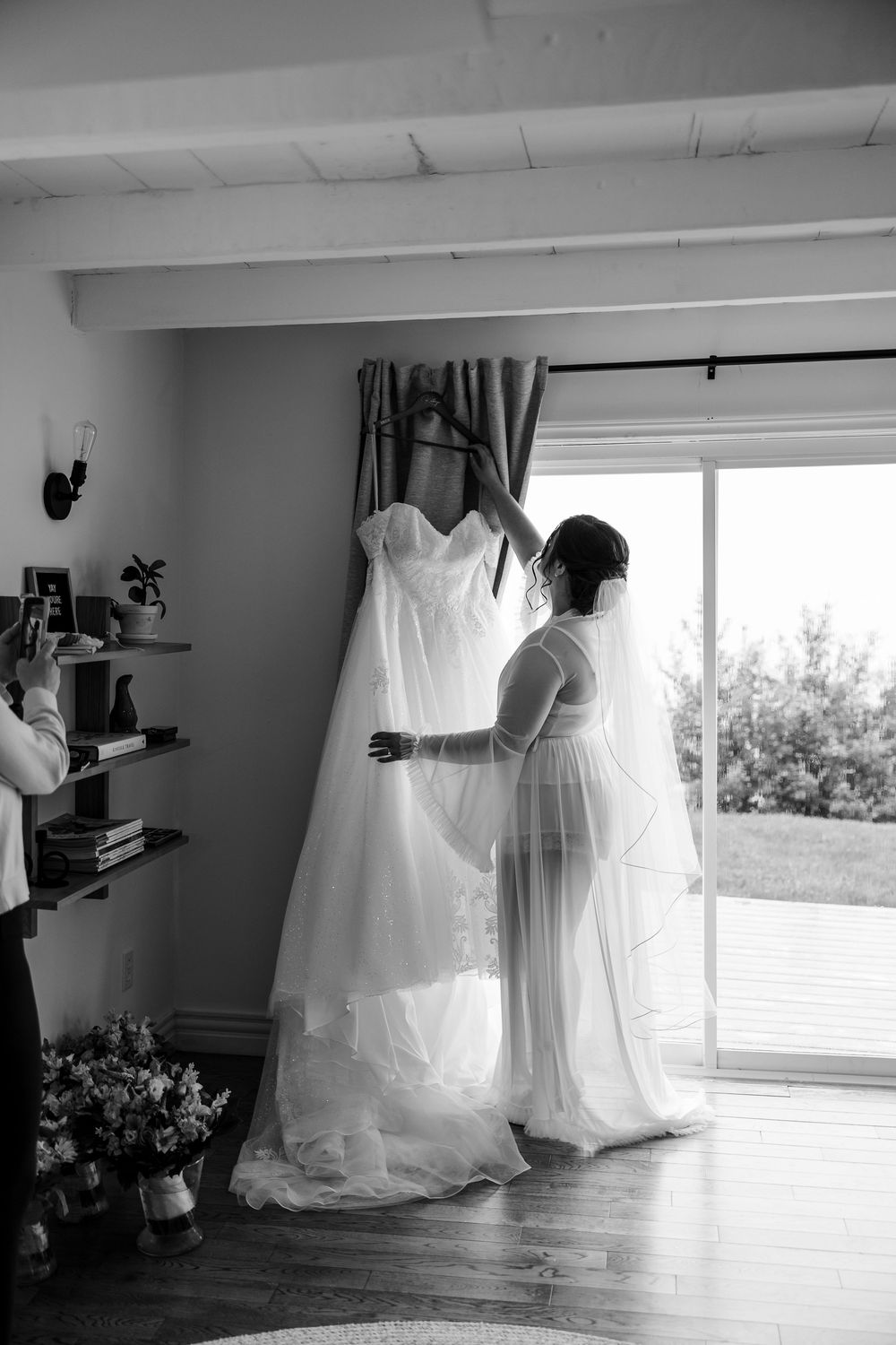 Black and white photo of a bride examining her wedding gown hanging by a sunlit window in a cozy room.