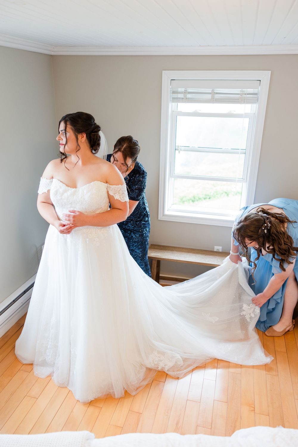 Bride in white off-shoulder gown being helped by two bridesmaids in blue dresses in a bright room.