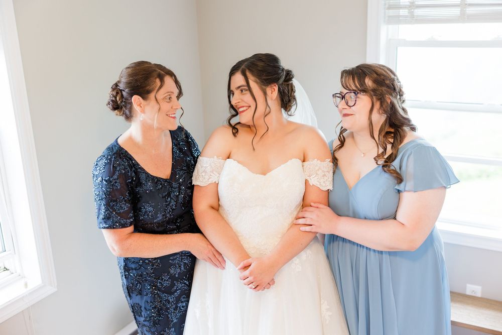 Bride in white off-shoulder gown smiles with two women in blue dresses during wedding preparations indoors.