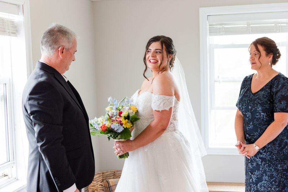 Bride in white off-shoulder gown holding colorful bouquet smiles at man in black suit during indoor wedding ceremony.