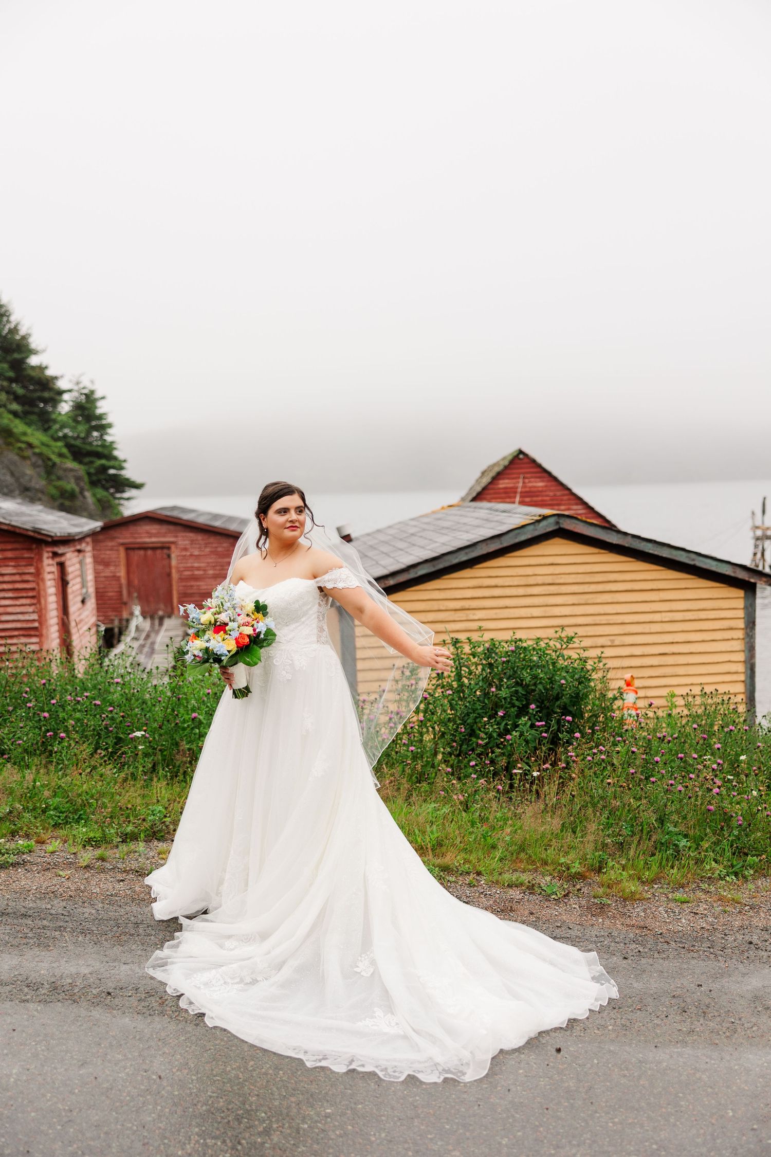 Bride in white off-shoulder gown with veil holds colorful bouquet outdoors near rustic buildings on overcast day.