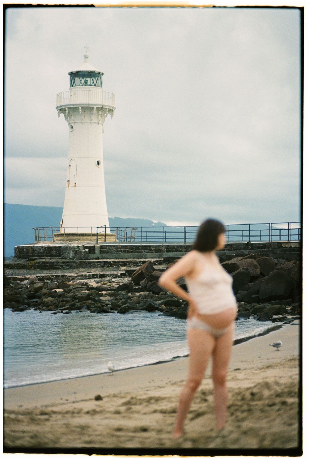 Woman in swimsuit standing on sandy beach gazing at a white lighthouse on rocky shore under cloudy skies.