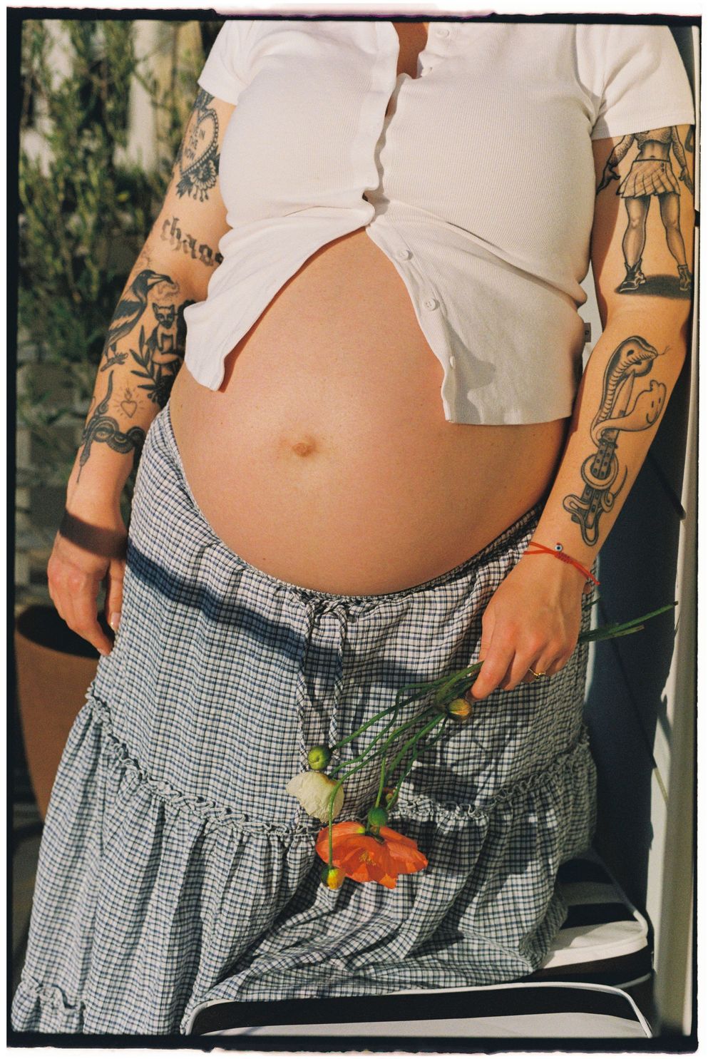 Pregnant woman with tattoos lifting white crop top to show baby bump, holding orange poppy flower in plaid skirt.