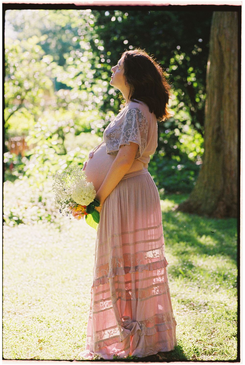 Pregnant woman in pink lace maxi dress holding flower bouquet, standing in sunlit garden during maternity photo session.