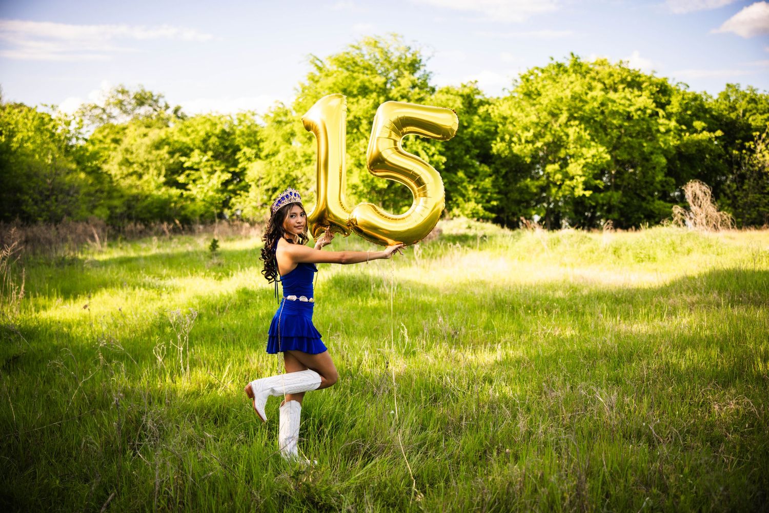 Girl in blue dress holds gold number 15 balloon in green field, celebrating her quinceañera outdoors.