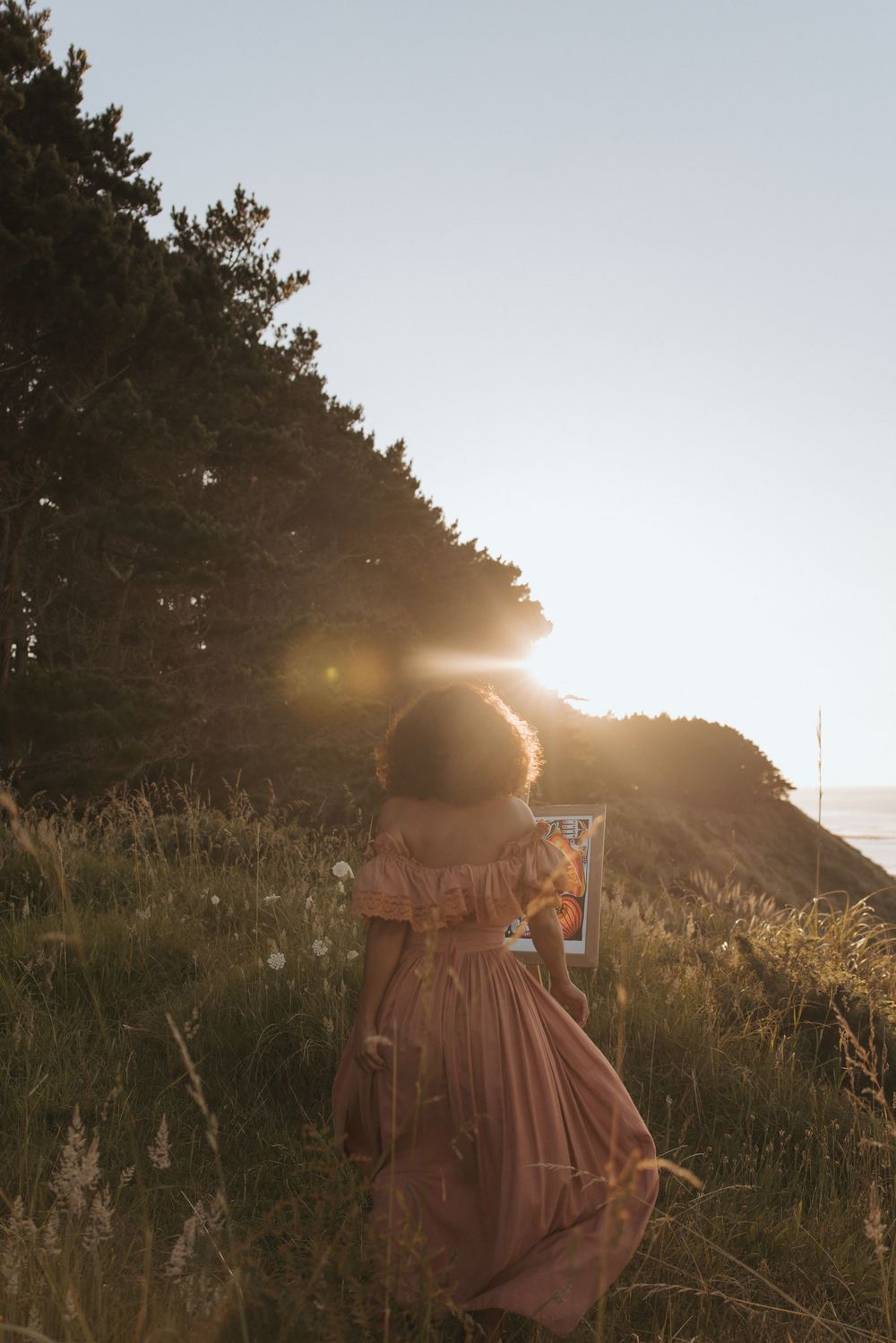Woman in pink off-shoulder dress walking through golden meadow at sunset with lens flare and trees.