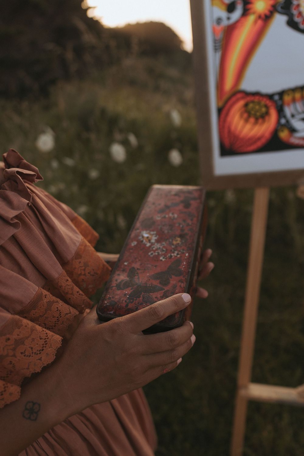 Woman in pink dress holding an ornate vintage book outdoors near an easel with colorful artwork at golden hour.