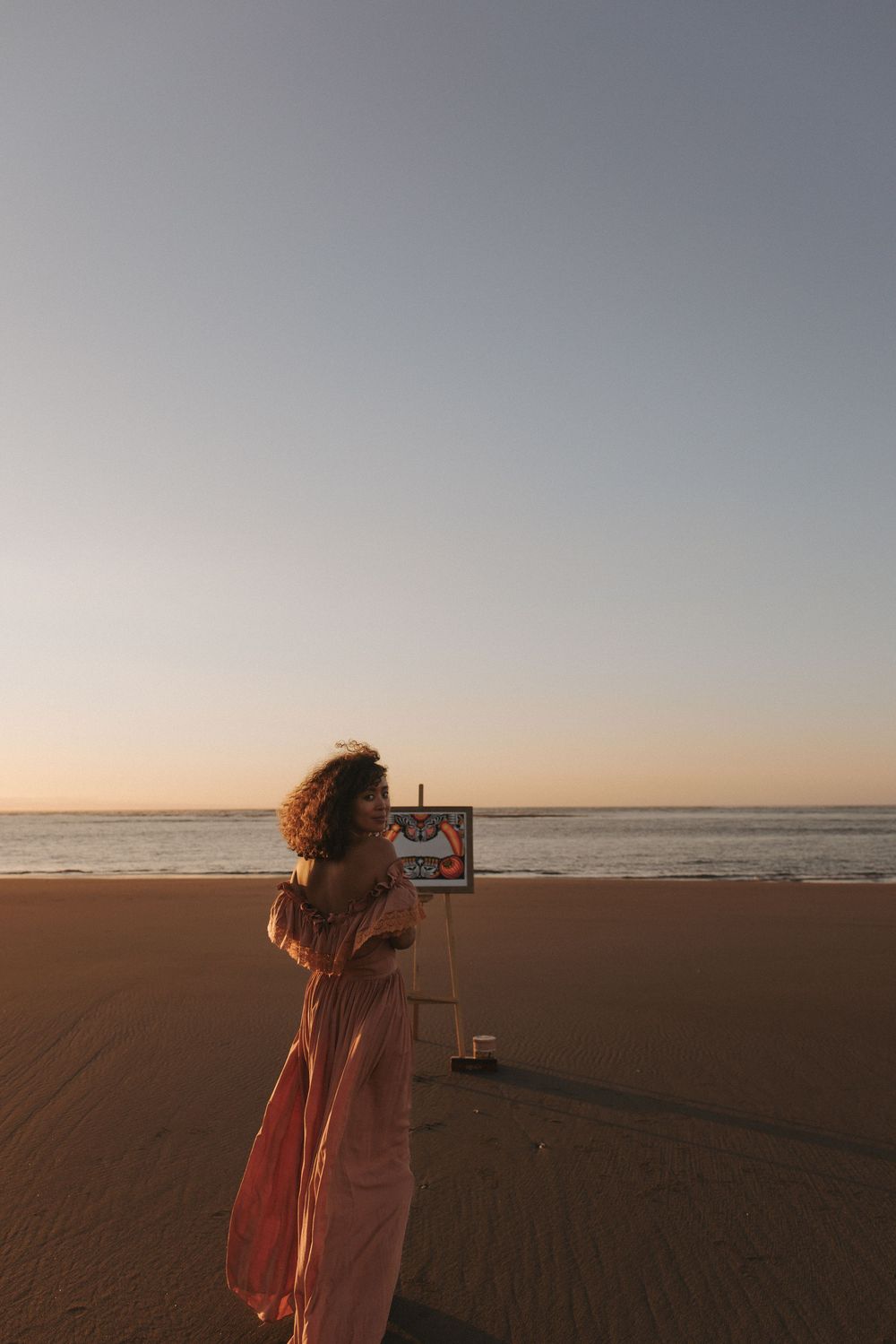 Woman in pink dress painting on easel at sunset beach, golden hour light reflecting on calm ocean waters.