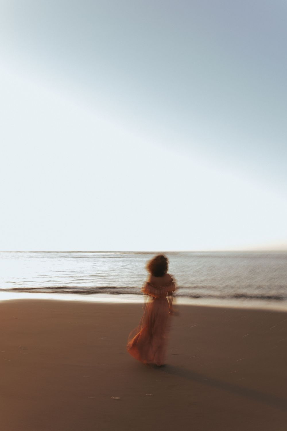 A blurred figure in a flowing orange dress stands on a sandy beach at sunset, gazing toward the calm ocean.