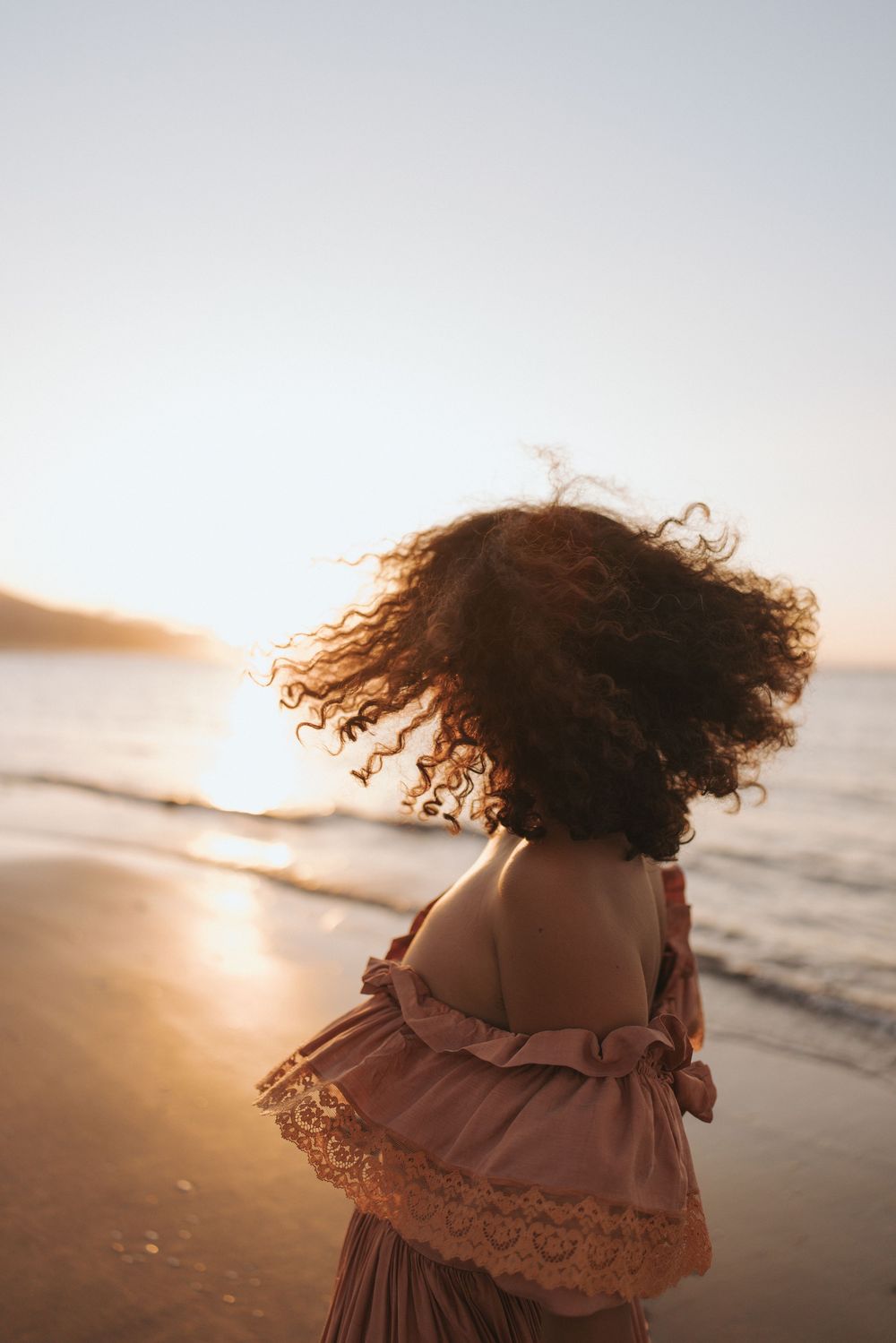 Woman in pink lace dress with curly hair blowing in wind on beach at golden sunset.