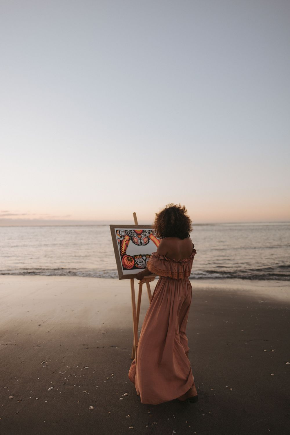 Woman in pink dress painting on easel at beach during sunset.