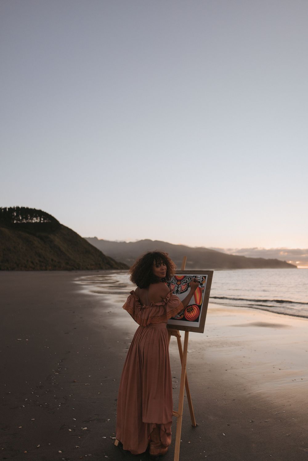 Woman in pink dress stands beside an easel with artwork on a sandy beach at sunset with hills in the background.