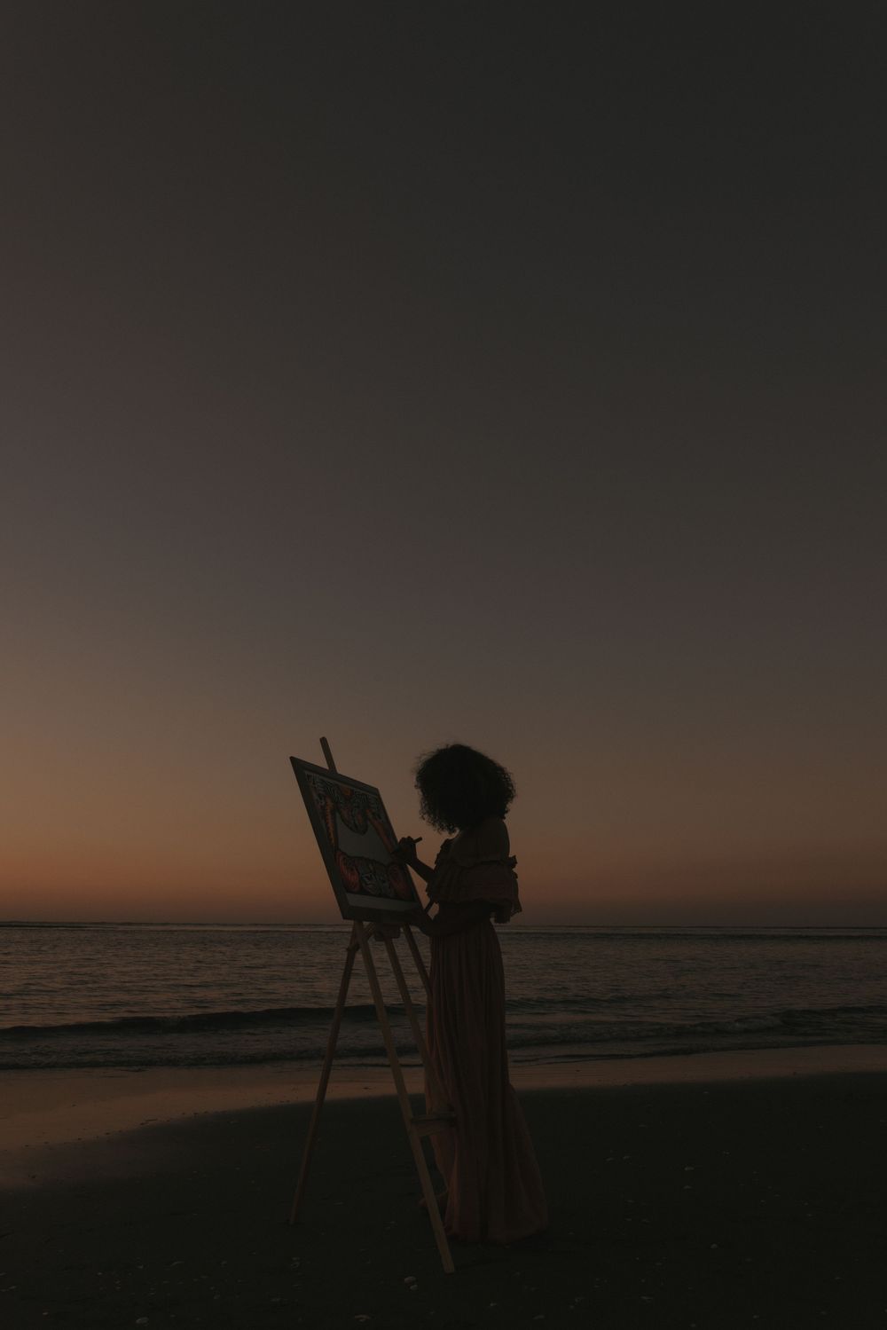 Silhouette of a woman painting on an easel at the beach during a stunning sunset with calm ocean waves.