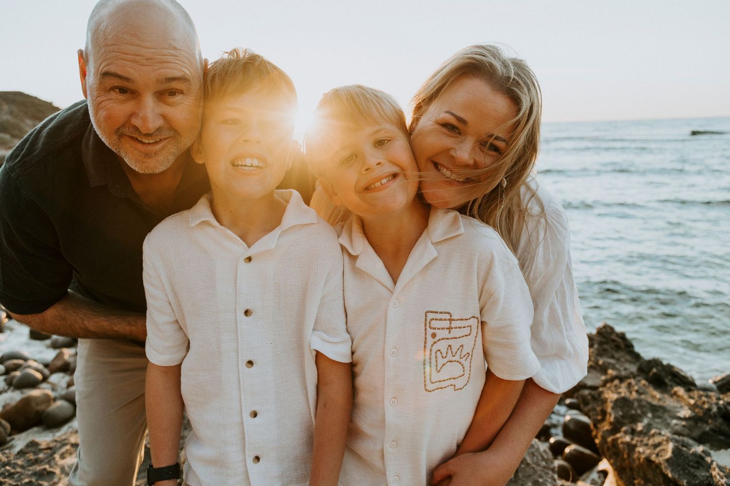 Family of four smiling together on rocky beach at golden sunset with ocean backdrop.