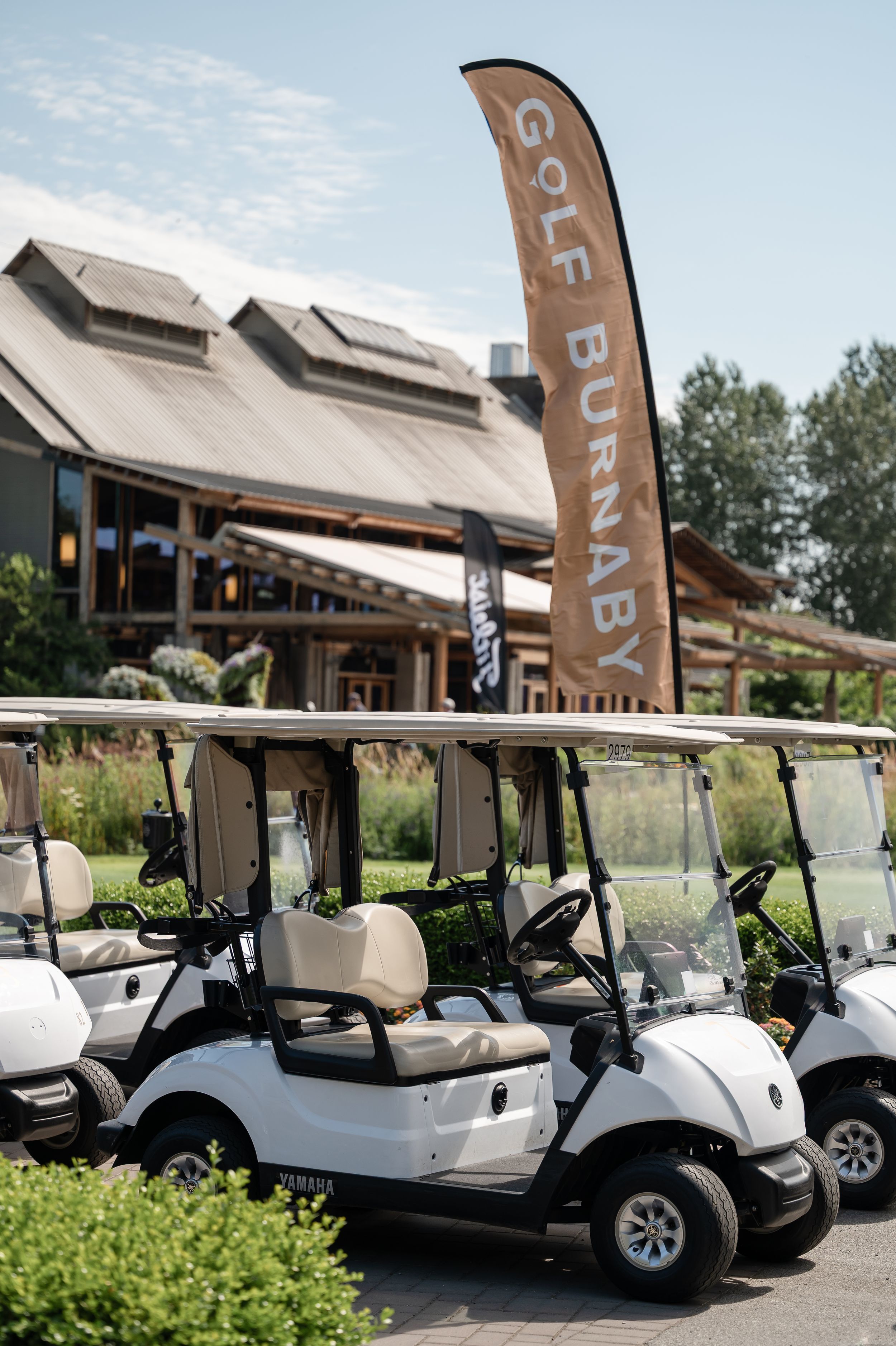 White Yamaha golf carts parked outside Golf Burnaby clubhouse beneath a tan feather flag on a sunny day.