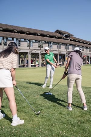 Three women practicing golf swings on a sunny green course near a clubhouse.