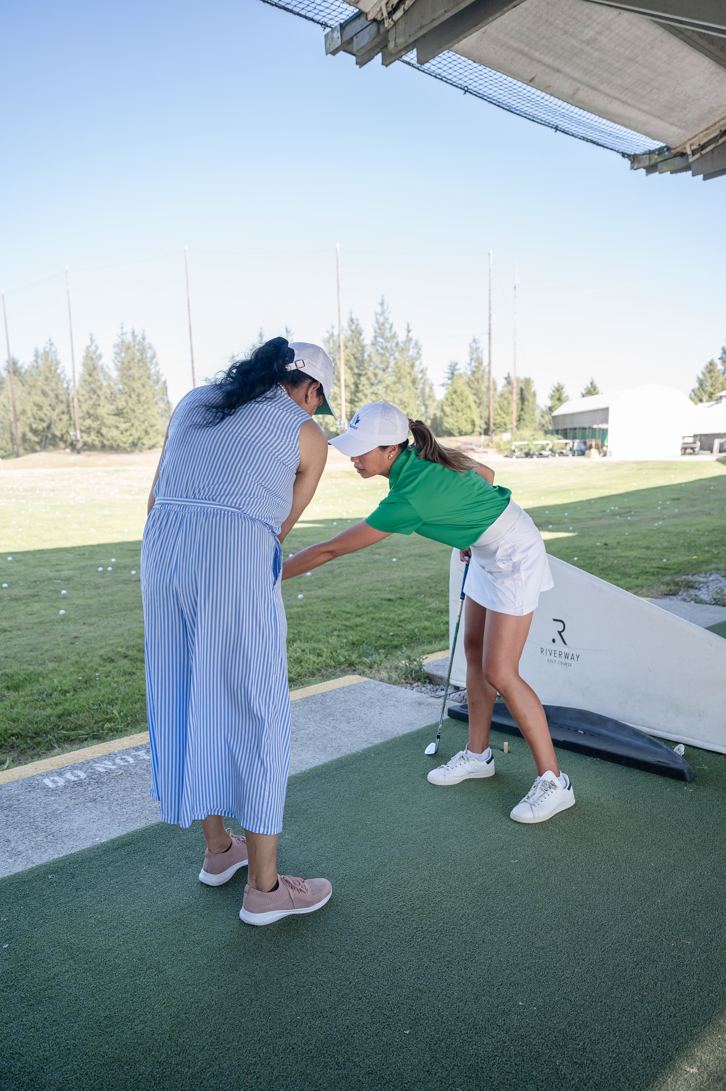 A golf instructor guides a young woman on her swing technique at a sunny outdoor driving range.