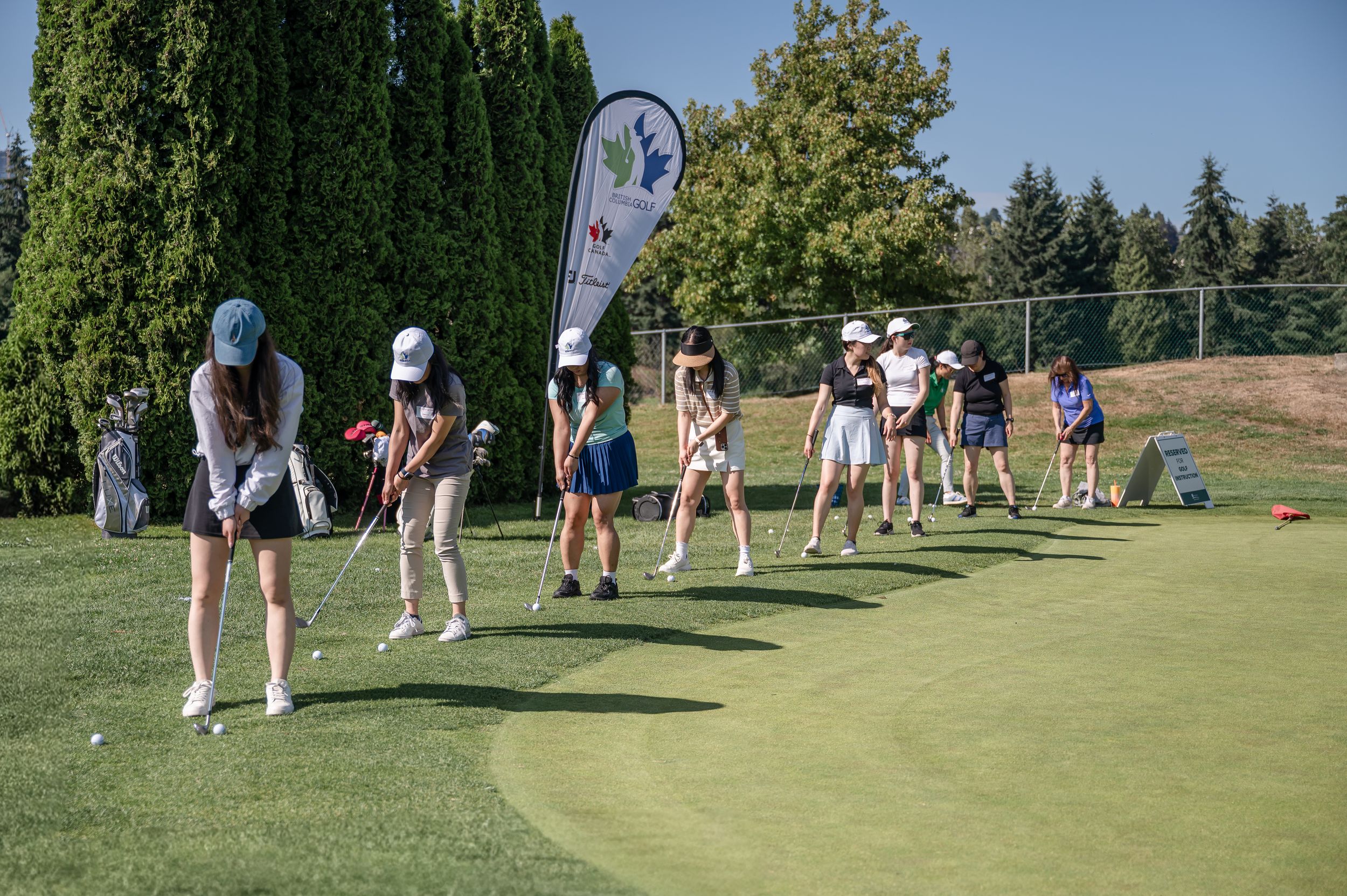 Young female golfers practice their swings on a sunny golf course driving range during a youth golf clinic.