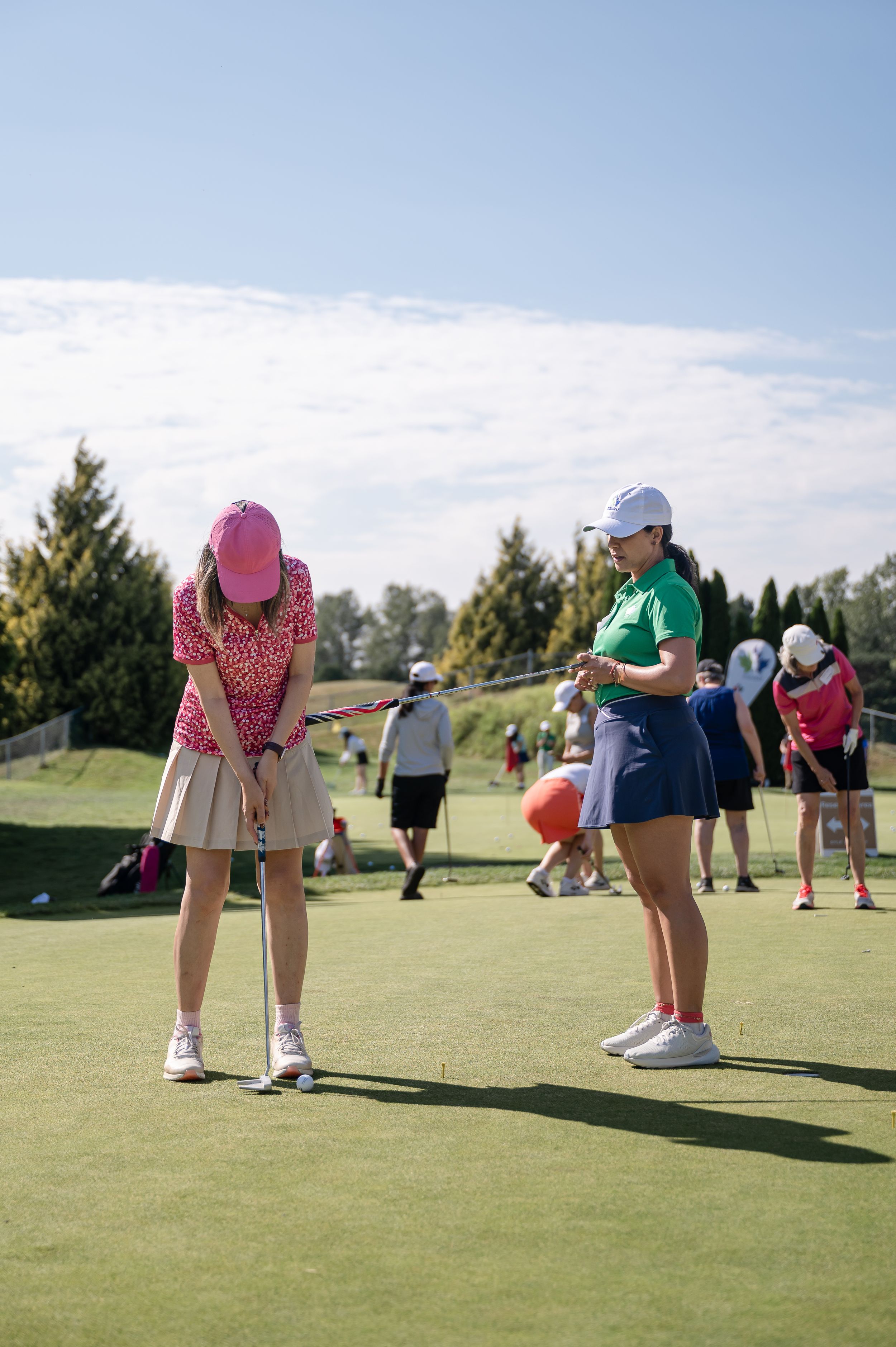 Two women practice putting on a golf green on a sunny day, one in pink and one in green attire.
