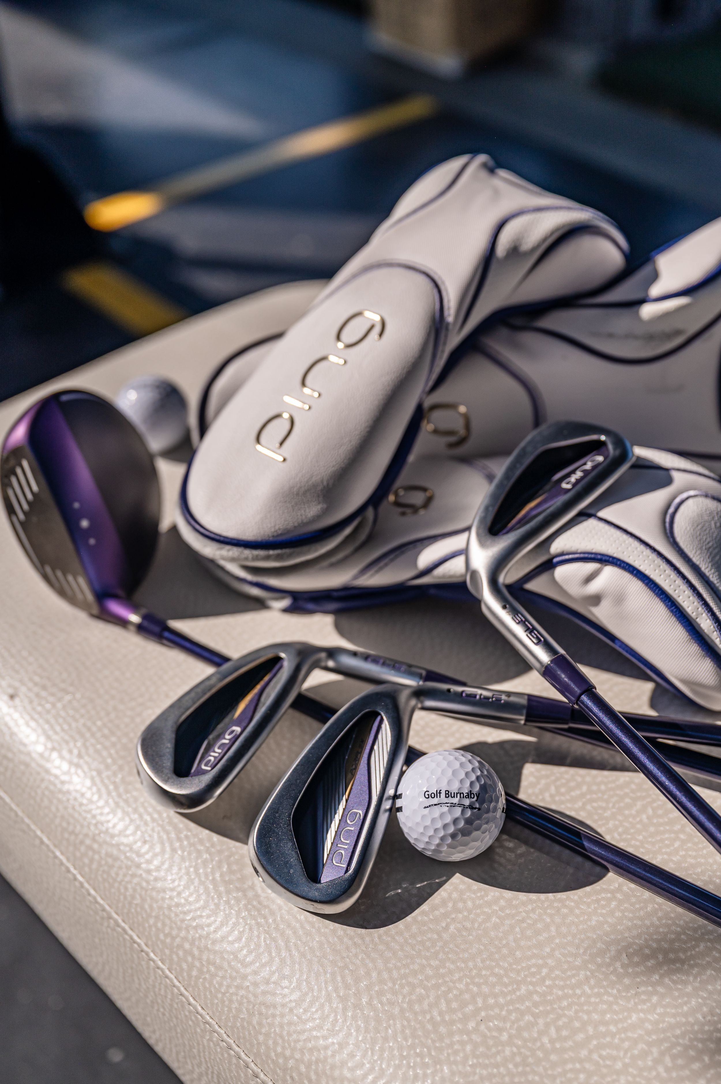 Ping golf clubs with purple accents and white headcovers rest beside a golf ball on a white golf bag.