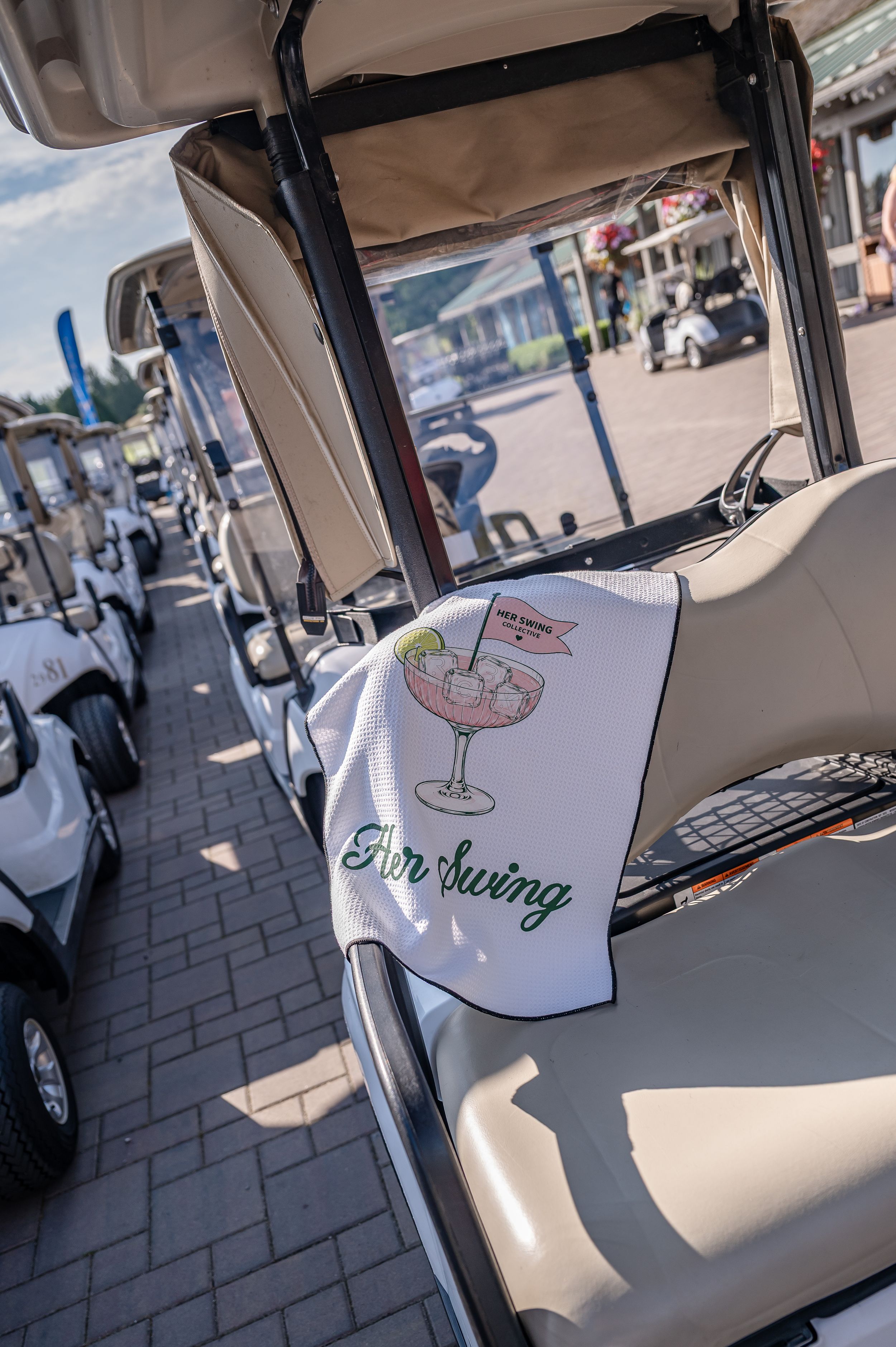 Golf cart with a decorative towel featuring a cocktail graphic and "Her Swing" text at a golf course event.