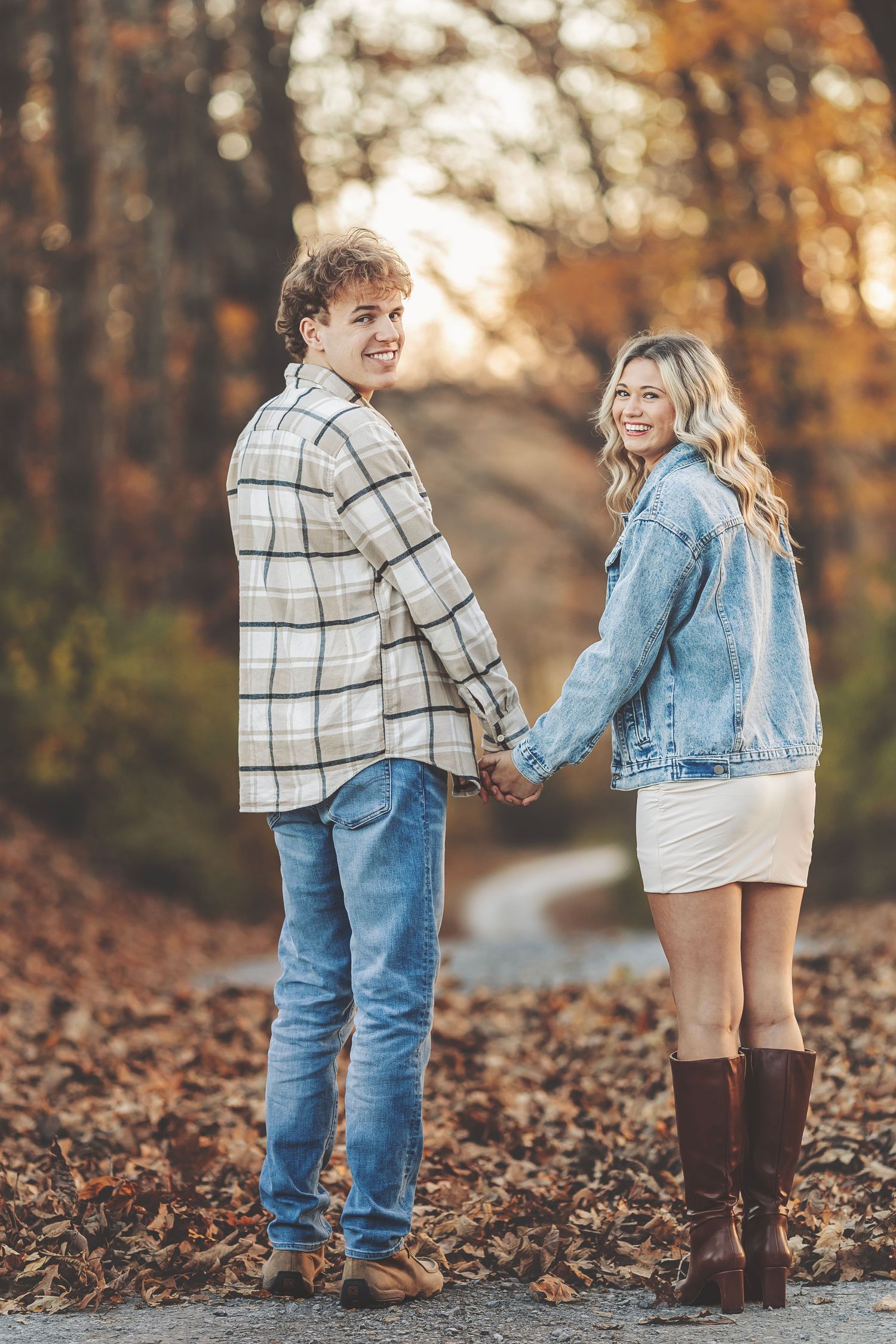 Couple holding hands on a leaf-covered path in autumn forest, wearing denim and plaid jackets, smiling over shoulders.