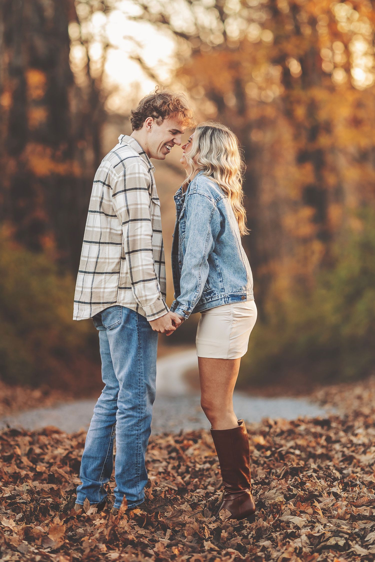 Couple holding hands and smiling at each other in a fall forest setting, wearing casual autumn outfits.